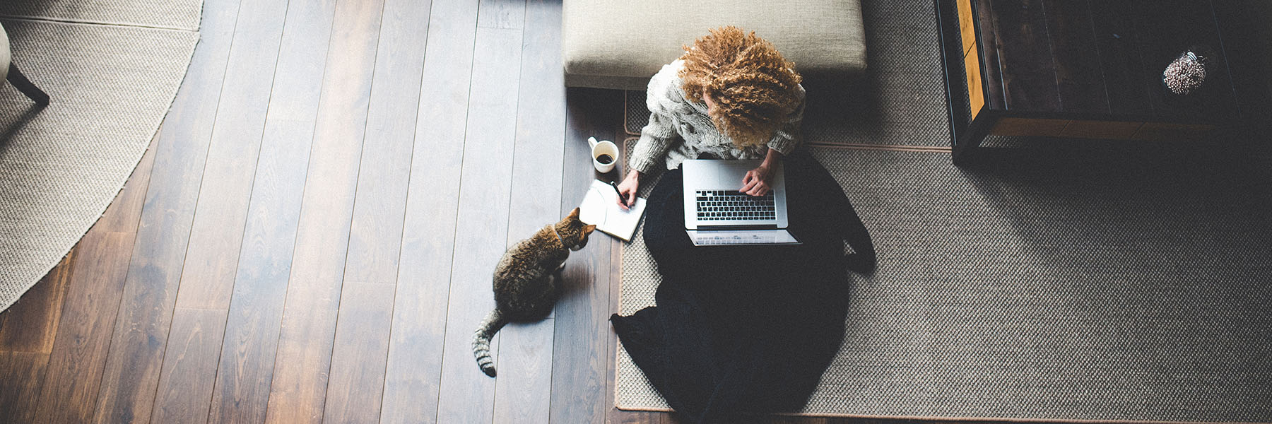 A woman sitting on the floor working on some papers. She has a cat watching her and a laptop on her lap.